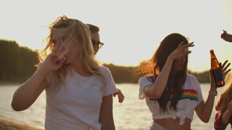 Tres-Estudiantes-Bailan-Con-Camisetas-Cortas-Con-Cerveza-En-La-Fiesta-En-La-Playa-Al-Atardecer.-Su-Largo-Cabello-Vuela-Con-El-Viento.-Esta-Es-Una-Tarde-De-Verano-Calurosa-Y-Despreocupada.